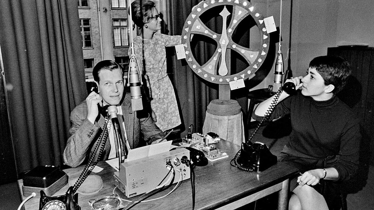 Sort-hvidt foto af tre personer, mikrofoner, telefoner og et lykkehjul i et radiostudie